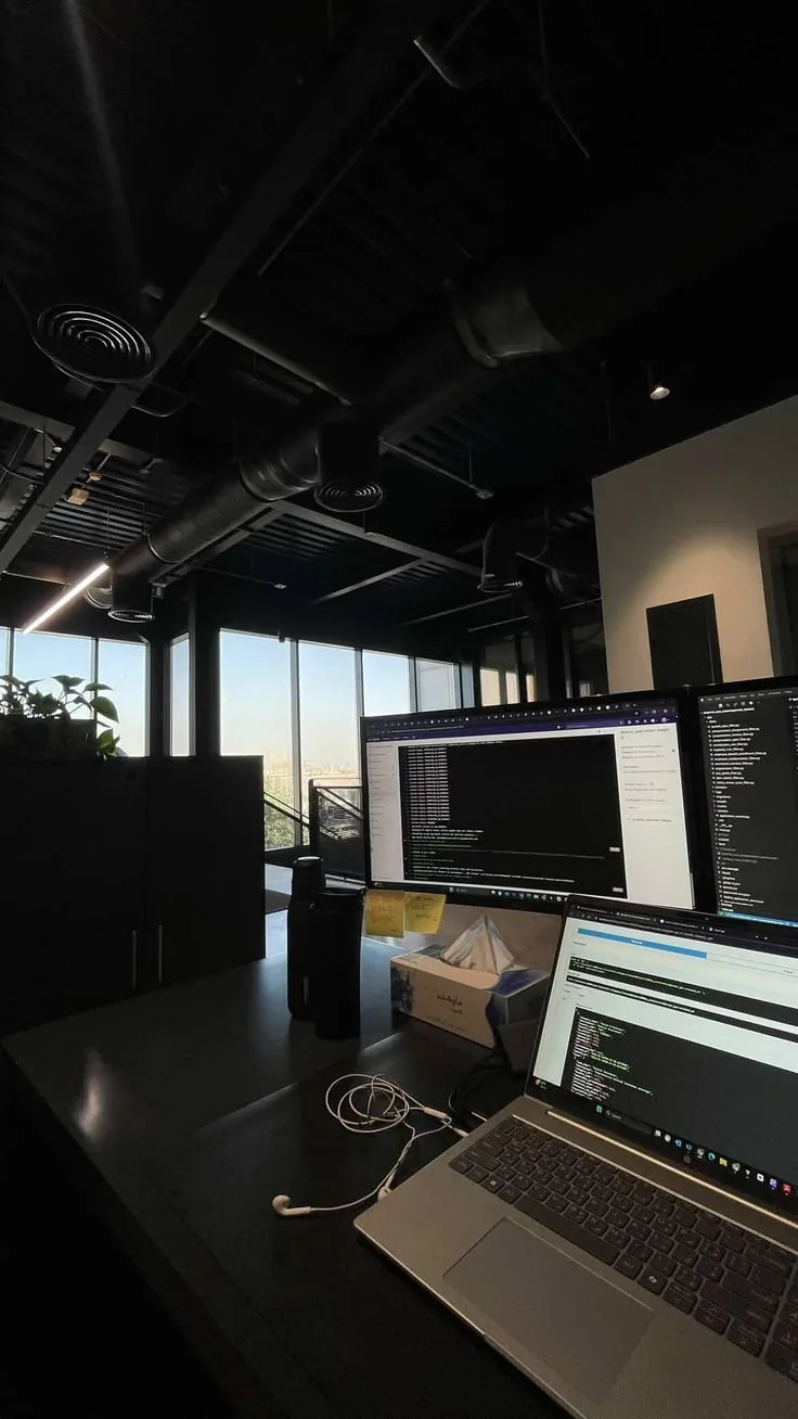 A studio desk with a laptop and two monitors, code on the screens, and daylight through the office windows.